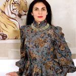 A photo of AGpress Founder Alessandra Guerrieri dressed in a suntuous metallic jacquard suit in charcoal and dark golden leaves, wearing a pearl chocker, standing majestically in front of a wall painted tiger