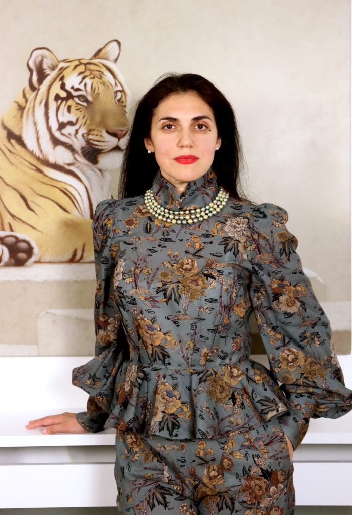 A photo of AGpress Founder Alessandra Guerrieri dressed in a suntuous metallic jacquard suit in charcoal and dark golden leaves, wearing a pearl chocker, standing majestically in front of a wall painted tiger
