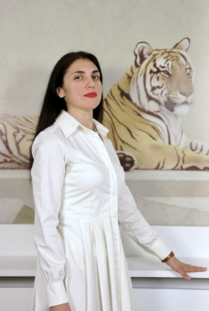 A photo of AGpress Founder Alessandra Guerrieri dressed in a tailored white shirt dress, standing majestically in front of a wall painted tiger
