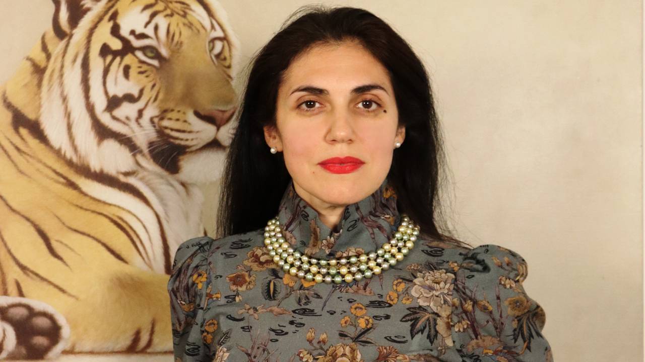 A photo of AGpress Founder Alessandra Guerrieri dressed in a suntuous metallic jacquard suit in charcoal and dark golden leaves, wearing a pearl chocker, standing majestically in front of a wall painted tiger